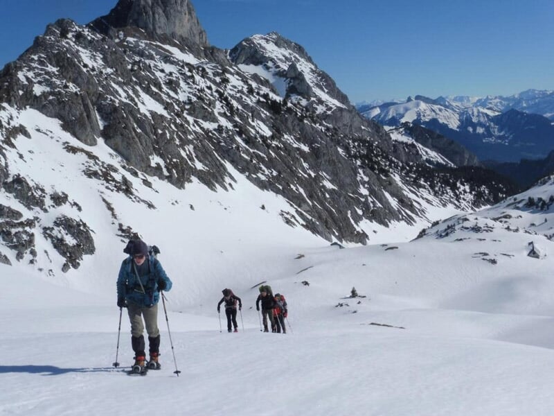 Randonneurs en raquettes progressant sur une pente enneigée avec des montagnes en arrière-plan.