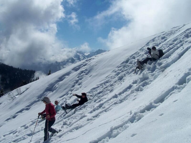 Personnes en raquettes descendant une pente enneigée en montagne sous un ciel partiellement nuageux.