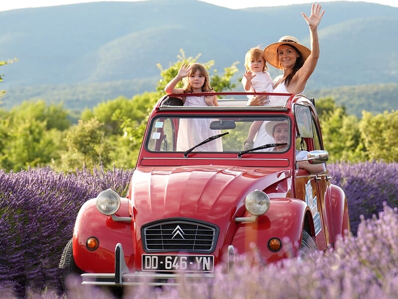 une Citroën 2CV rouge avec un homme au volant et une femme et deux enfants saluant depuis le toit ouvrant, stationnée dans un champ de lavande, avec des montagnes et un ciel d'été en arrière-plan