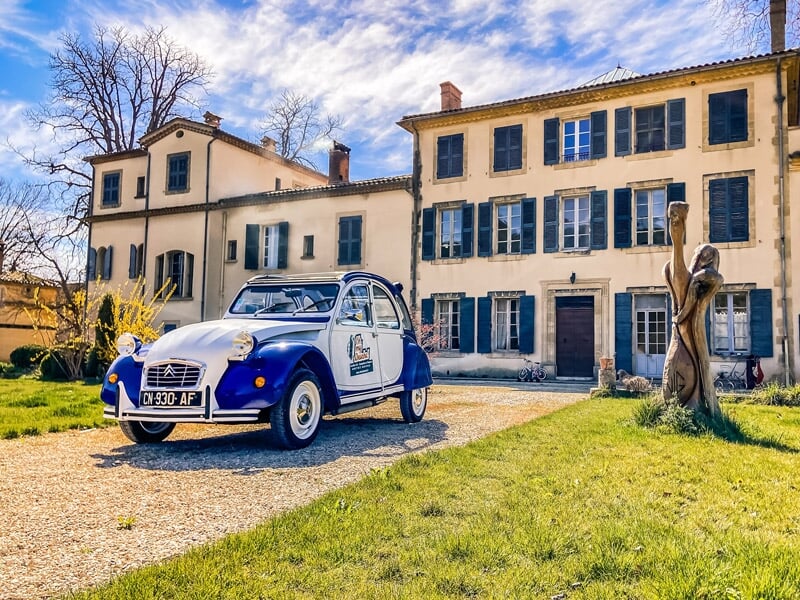 Citroën 2CV bleue et blanche avec des autocollants publicitaires, garée devant une maison de campagne traditionnelle aux volets verts, sous un ciel bleu clair. Présence d'une sculpture en bois à droit