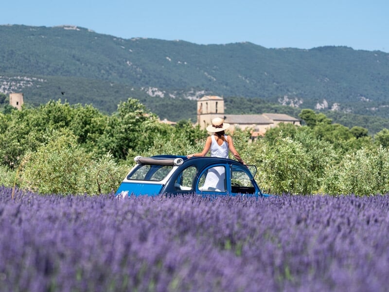 une femme portant un chapeau de paille et une robe blanche, debout à travers le toit ouvrant d'une Citroën 2CV bleue, au milieu d'un champ de lavande en fleur avec en arrière-plan un village et des mo