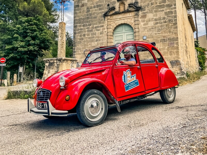 Vue dynamique d'une Citroën 2CV rouge, en mouvement sur une route de village, avec un homme au volant portant des lunettes de soleil et un passager souriant à travers la fenêtre ouverte, près d'un bât