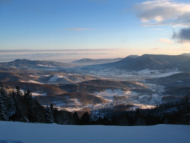 Vue panoramique d'une vallée montagneuse avec forêts et champs sous un ciel bleu clair et lumineux.
