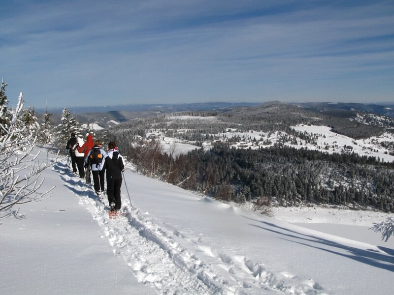 Randonneurs en raquettes marchant sur un sentier enneigé avec vue panoramique sur la vallée et montagnes.