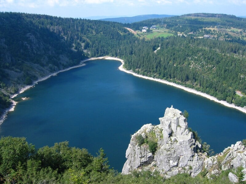 Vue aérienne d'un lac entouré de forêts vertes et collines sous un ciel clair en été.