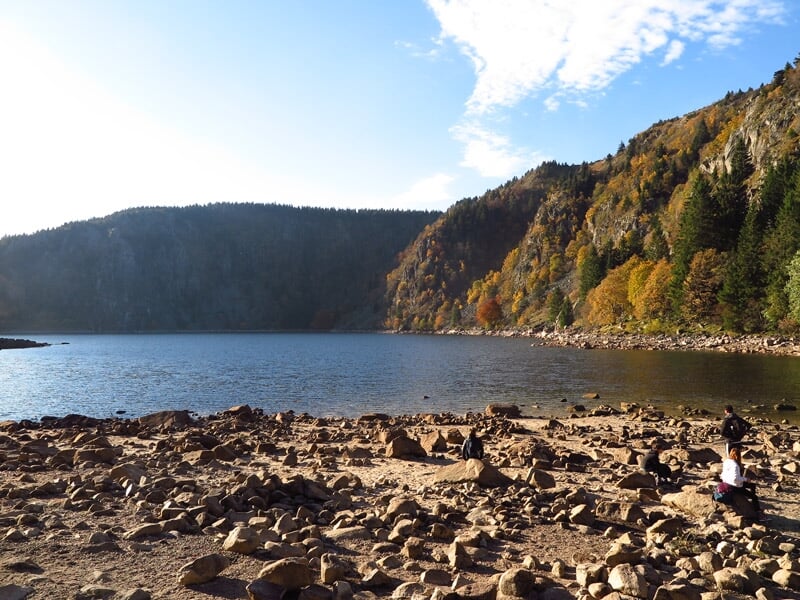 Bord d'un lac avec eau calme, rochers au premier plan et forêt colorée en automne sous un ciel bleu.