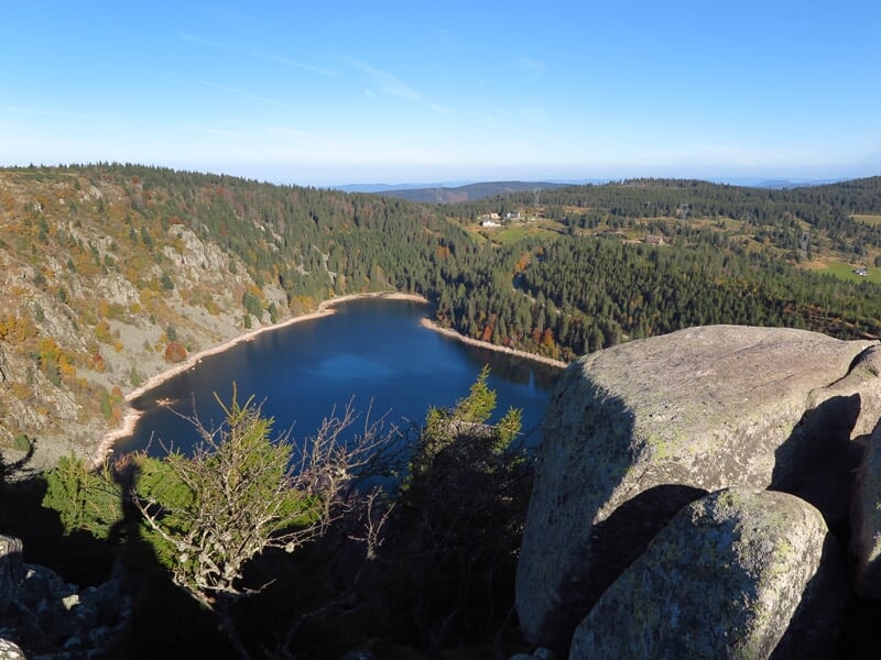 Vue panoramique d'un lac entouré de collines boisées avec un grand rocher au premier plan.