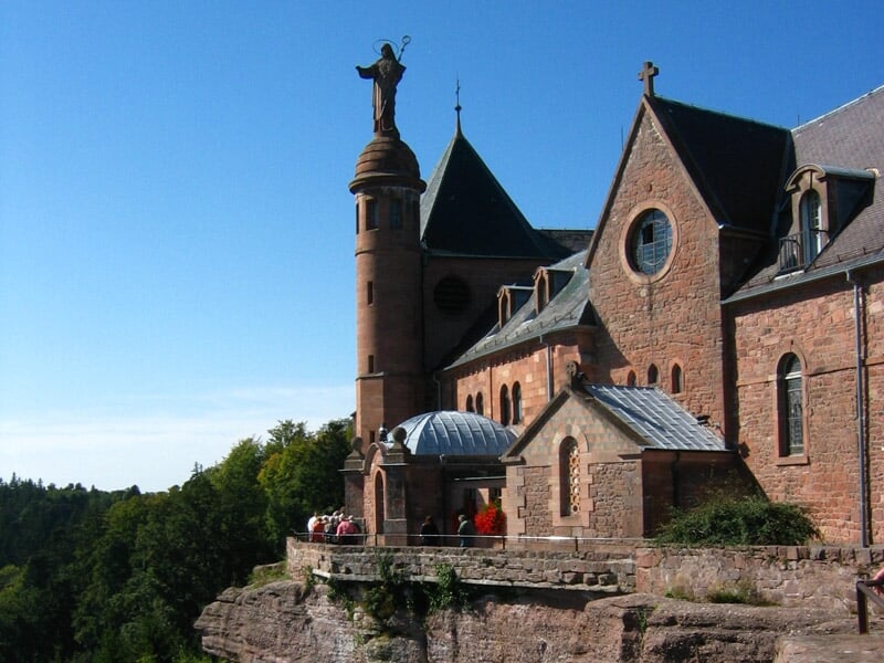 Église ou abbaye en pierre rouge avec statue sur une colline boisée sous un ciel clair