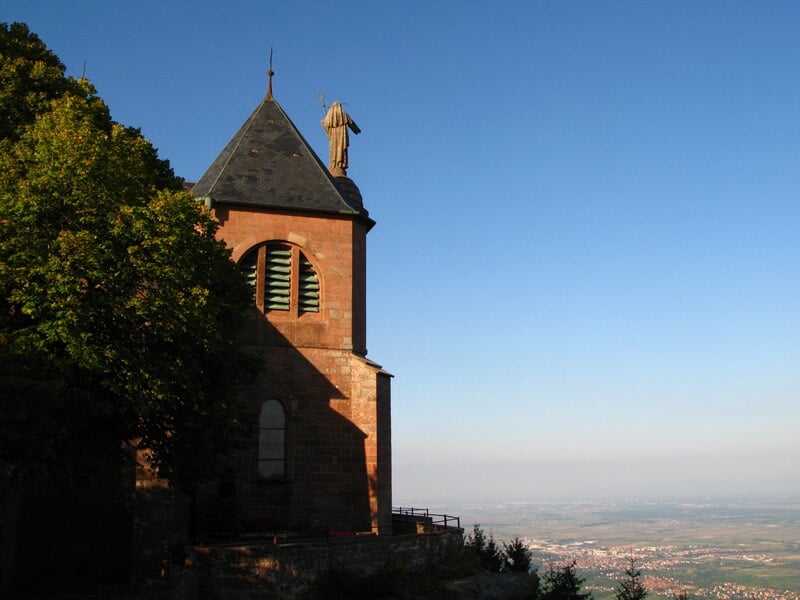 Église en pierre rouge avec clocher et arbres, vue sur la plaine en arrière-plan