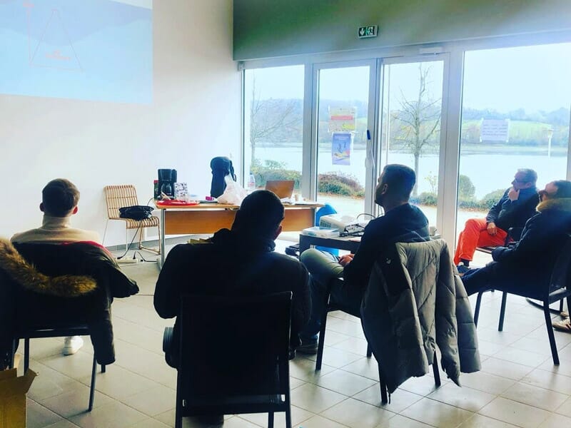 Groupe de personnes assises en salle, regardant une présentation avec vue sur le lac à travers les fenêtres.