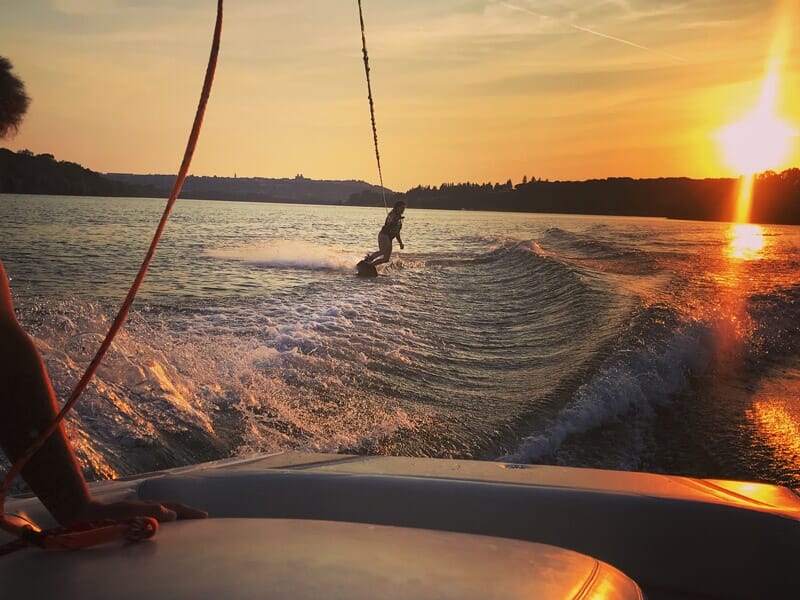Personne pratiquant le wakeboard au coucher du soleil sur le lac de la Liez, avec des vagues derrière le bateau.