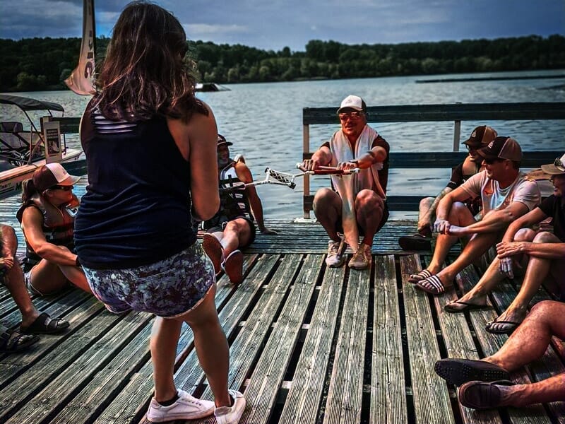 Groupe de personnes assises sur un ponton en bois au bord du lac, discutant et profitant du soleil.