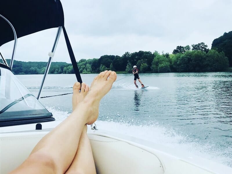 Personne allongée sur un bateau, jambes étendues, regardant quelqu'un faire du wakeboard sur le lac.