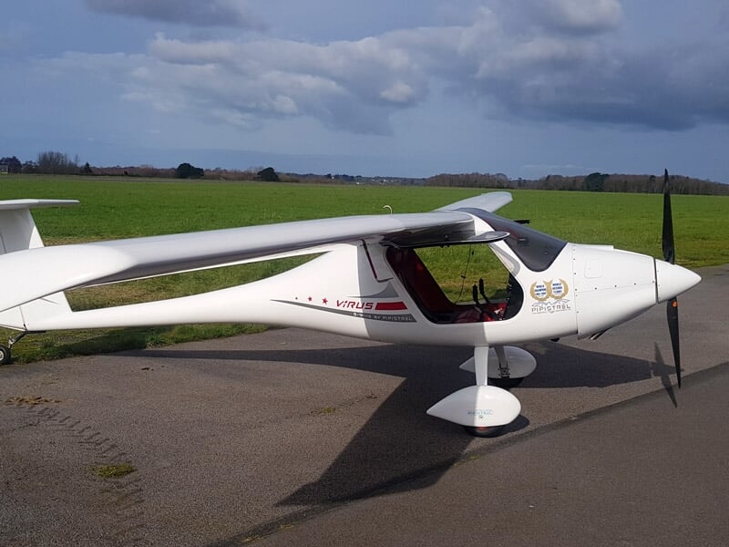 Vue latérale d'un avion léger blanc sur une piste, avec un champ vert et un ciel nuageux.