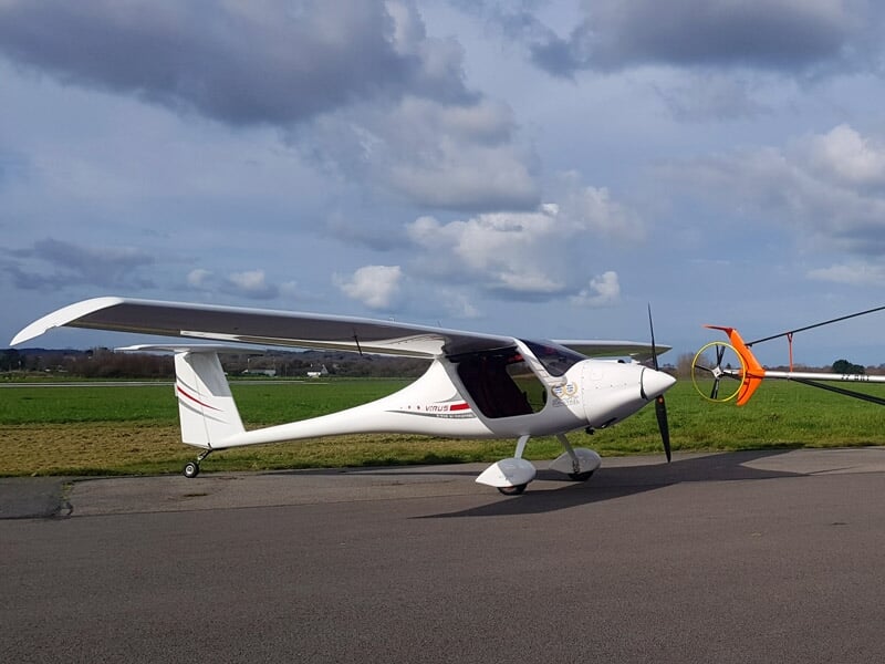 Avion léger blanc sur piste avec hélice orange à l'avant et ciel nuageux en arrière-plan.