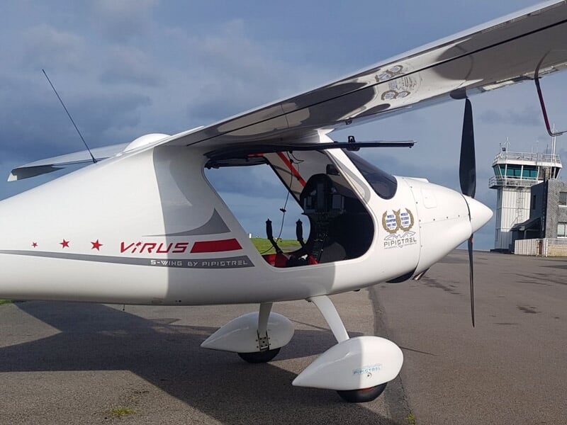 Vue latérale d'un avion léger blanc avec marquage Virus, stationné près d'un bâtiment d'aérodrome.