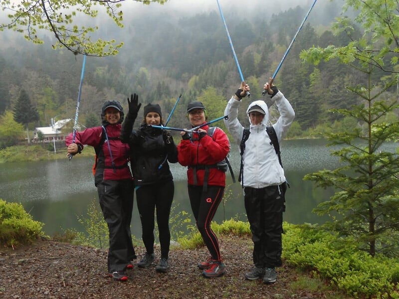 Quatre femmes souriantes avec bâtons de marche nordique posant près d'un lac en forêt.