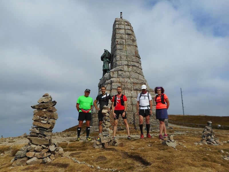 Groupe de cinq coureurs posant devant un monument en pierre au sommet d'une montagne.