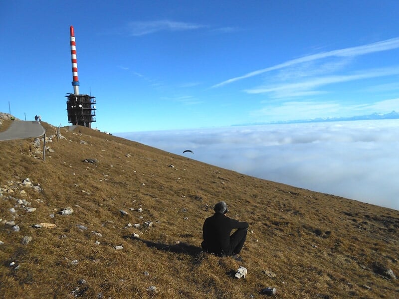 Personne assise sur une colline herbeuse regardant un paysage de montagnes et de nuages.