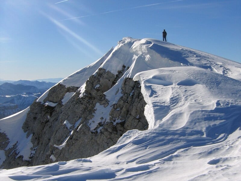 Personne debout au sommet d'une crête enneigée avec un ciel bleu clair et des montagnes en arrière-plan.