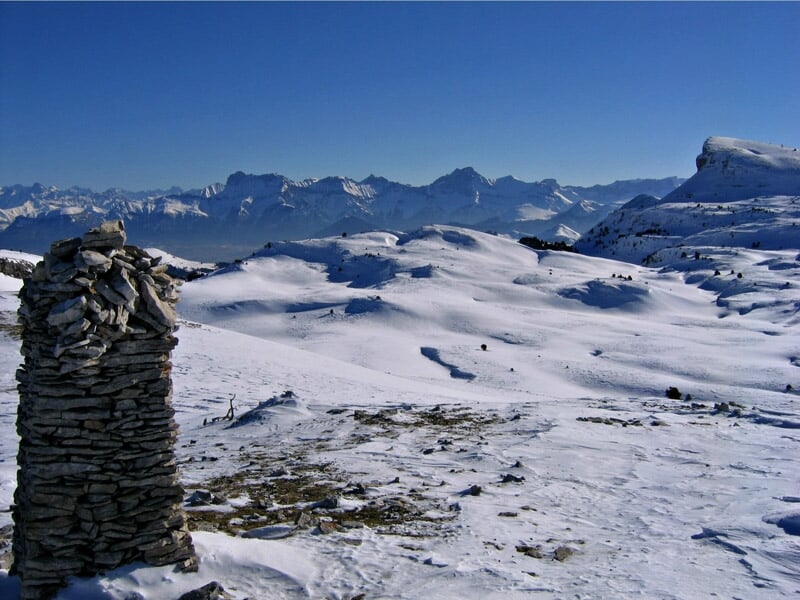 Paysage de montagne enneigée avec un cairn en pierre au premier plan et des sommets à l'horizon.