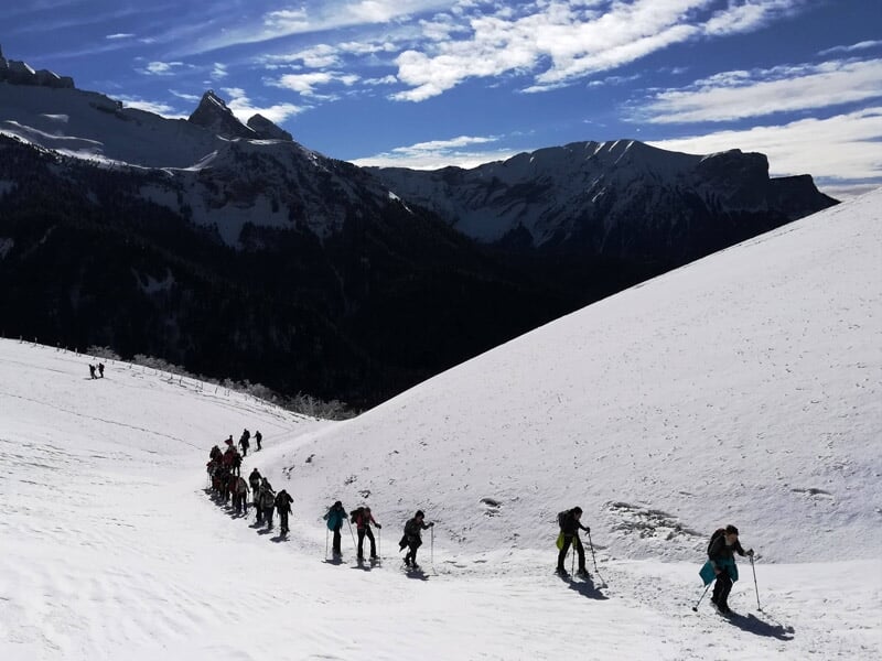 File de randonneurs en raquettes à neige progressant sur un sentier en pente dans un paysage montagneux.