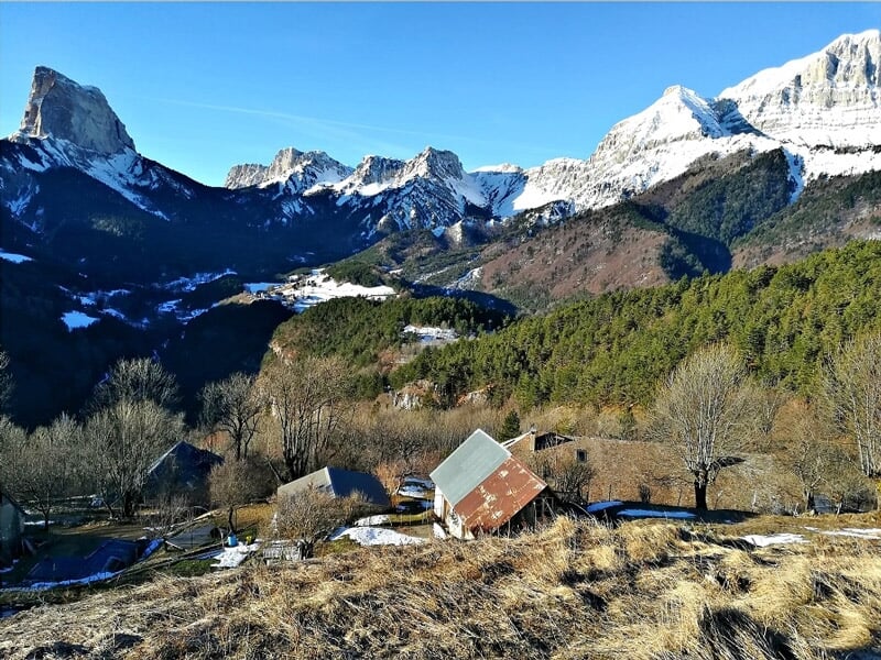 Village de montagne avec des maisons et des arbres au pied de sommets enneigés sous un ciel clair.