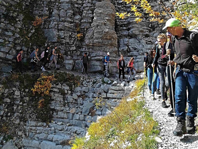 Groupe de randonneurs marchant sur un sentier étroit au pied d'une falaise rocheuse avec des feuilles d'automne.