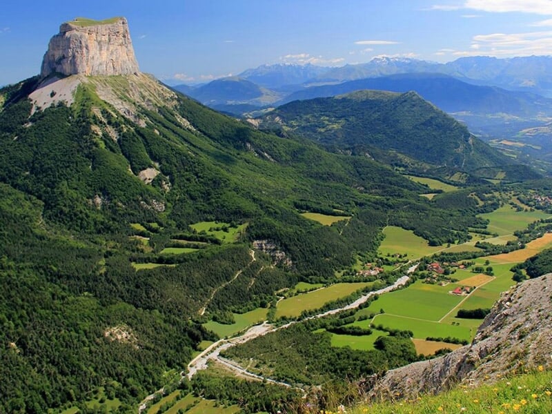 Vue panoramique d'une vallée verdoyante entourée de montagnes avec un sommet rocheux emblématique.