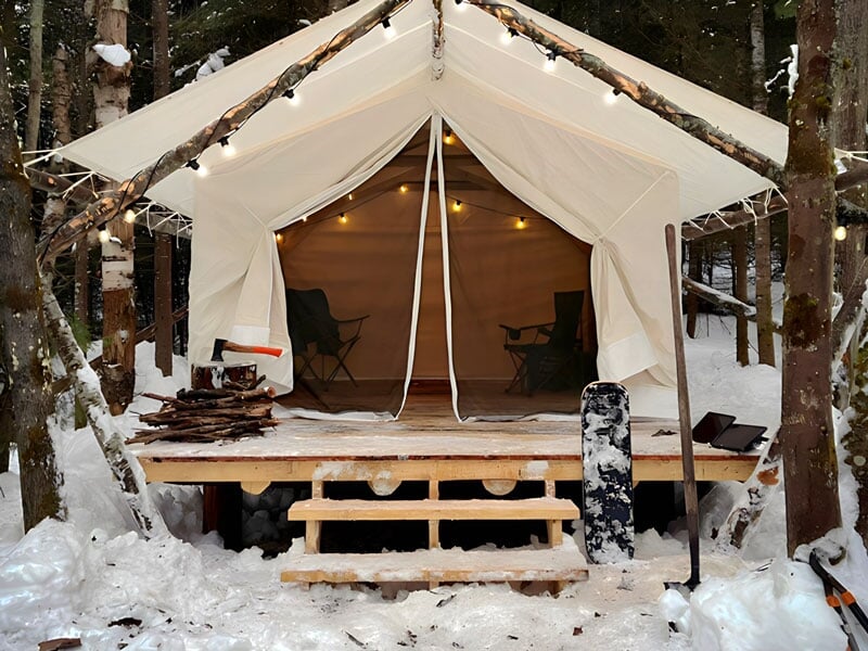 Tente blanche avec éclairage intérieur doux montée sur plateforme en bois dans un environnement enneigé entouré d'arbres, avec hache et bois de chauffage à l'avant