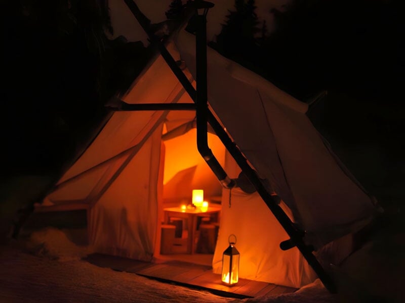 Tente de camping illuminée par une lumière chaude à l'intérieur et lanternes à l'extérieur, dans la forêt enneigée durant la nuit, évoquant une atmosphère chaleureuse