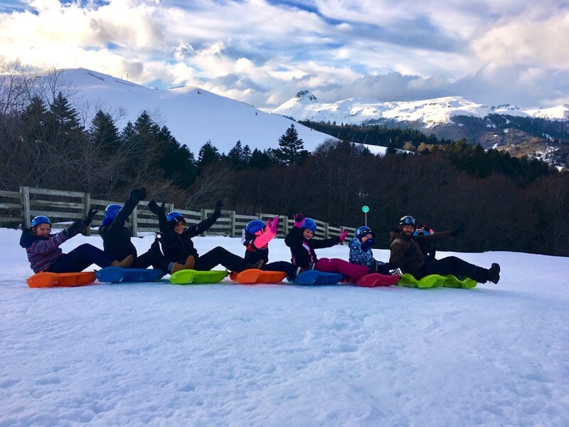 Groupe d'enfants assis sur des luges colorées alignées sur la neige, portant casques et vêtements d'hiver, avec montagnes en arrière-plan, signifiant divertissement hivernal