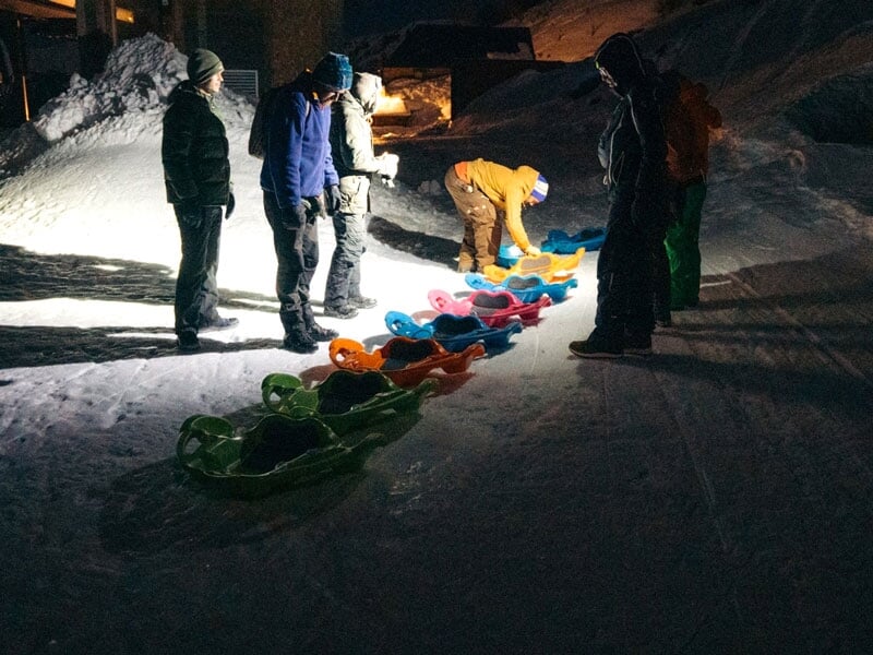 Personnes préparant des luges sur une pente enneigée éclairée de nuit, vêtues de tenues de neige et de bonnets, suggérant des activités de glisse nocturnes