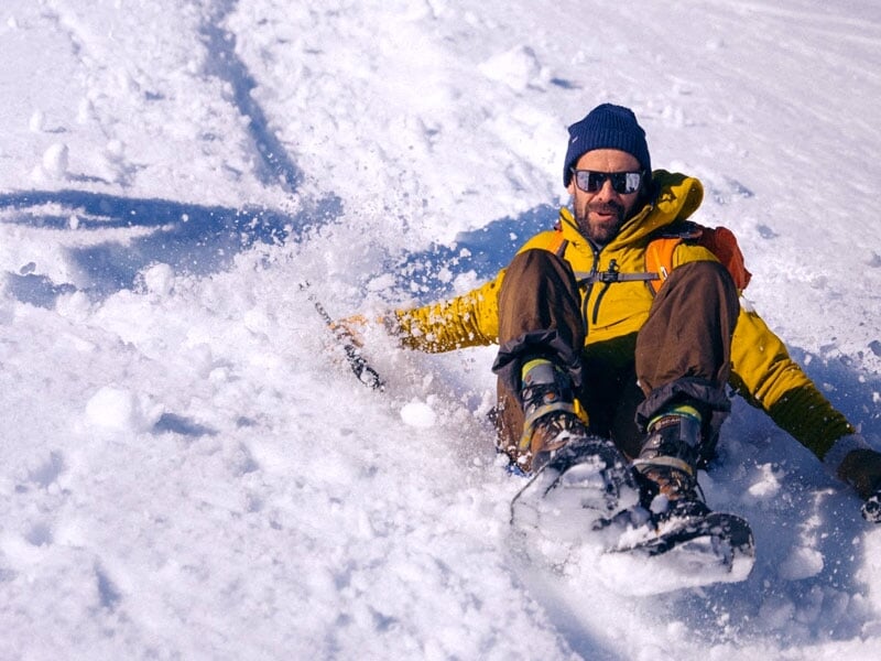 Homme glissant sur la neige en tenue de montagne avec sac à dos, lunettes de soleil et bonnet, laissant une traînée derrière lui, représentant les loisirs d'hiver actifs