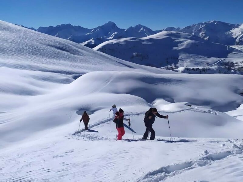 Quatre personnes en raquettes marchant sur une pente enneigée avec des montagnes en arrière-plan sous un ciel bleu clair.