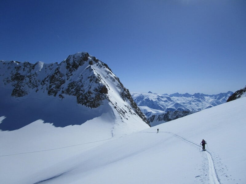 Deux randonneurs en raquettes gravissant une montagne enneigée avec un ciel bleu et des sommets alpins à l'horizon.