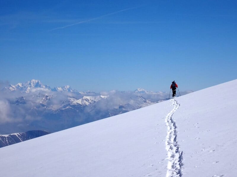 Personne seule en raquettes suivant une trace dans la neige sur une pente douce avec un ciel bleu et des montagnes lointaines.
