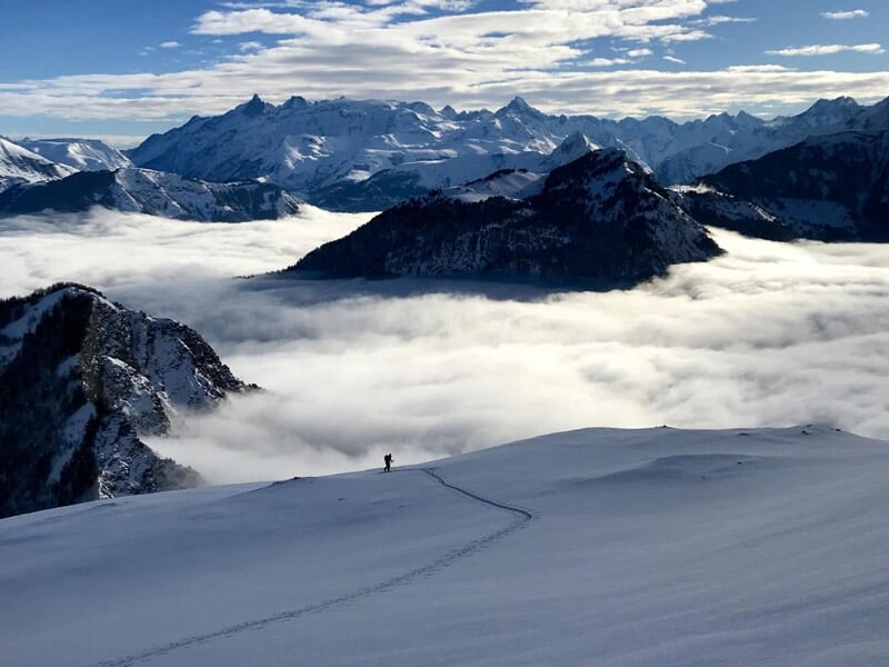 Vue panoramique d'une montagne enneigée avec un sentier tracé et un ciel partiellement nuageux au-dessus d'une mer de nuages.