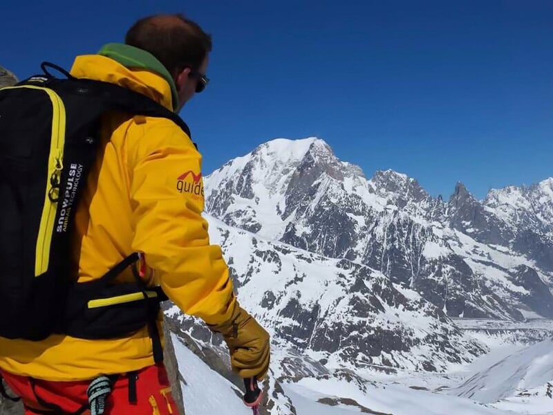 Guide en tenue jaune avec sac à dos observant un paysage montagneux enneigé sous un ciel bleu clair, prêt pour la randonnée.