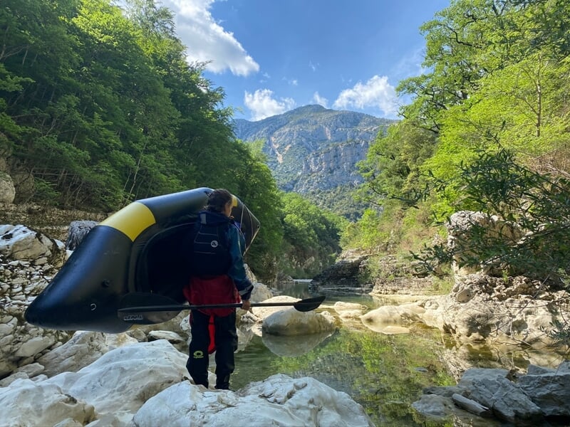 Personne portant un kayak gonflable noir et jaune près d'une rivière entourée de rochers et de montagnes verdoyantes.
