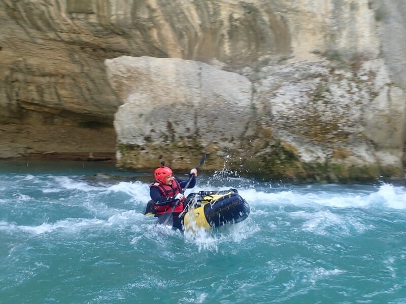 Kayakiste en casque rouge pagayant dans une eau vive turquoise au pied d'une falaise rocheuse imposante.