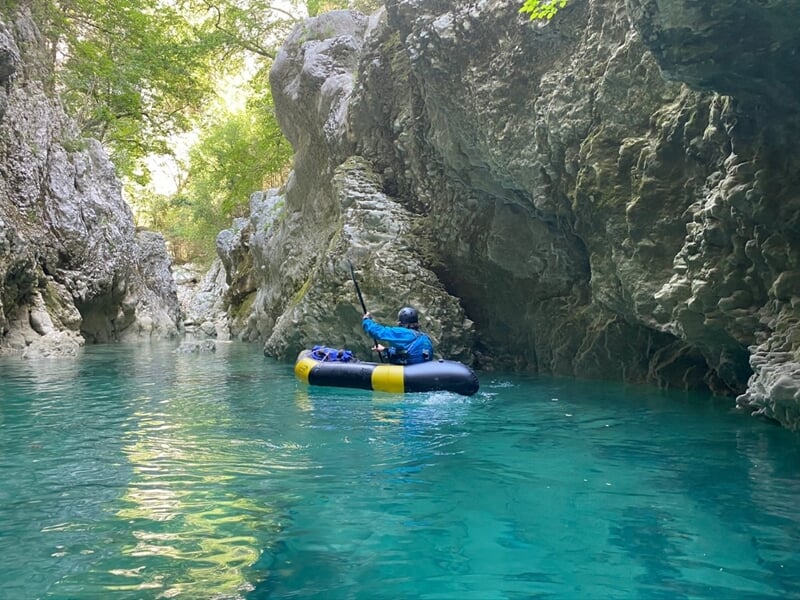 Individu en kayak gonflable bleu et jaune pagayant dans une rivière calme aux eaux claires et aux falaises rocheuses.