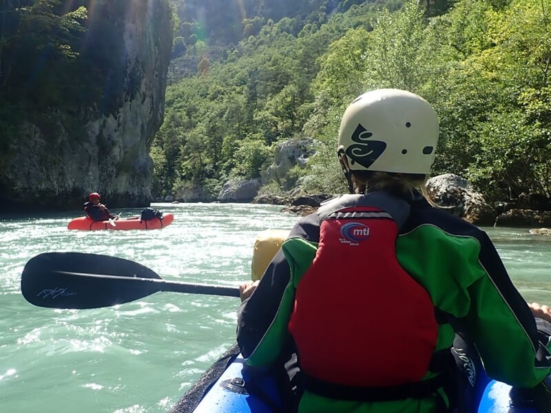 Vue arrière d'un kayakiste en casque blanc et gilet rouge pagayant sur une rivière entourée de falaises et de végétation.