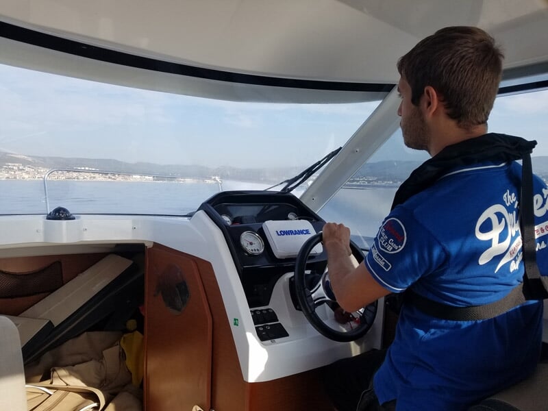 Un homme pilote un bateau à moteur en mer, avec la côte visible à l'horizon par une journée claire.