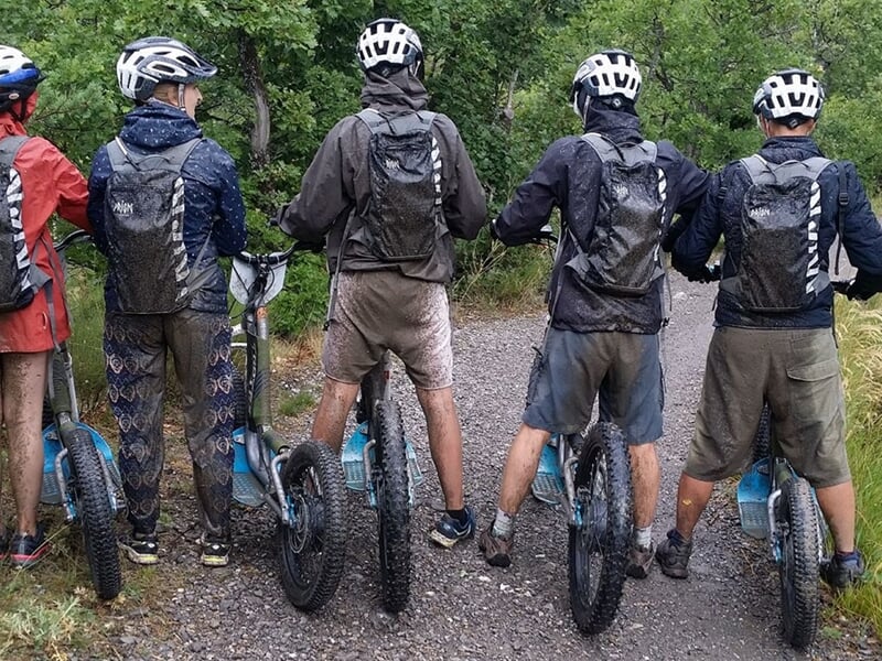Groupe de cinq personnes de dos avec casques et sacs à dos, prêtes à rouler sur des trottinettes électriques en forêt.