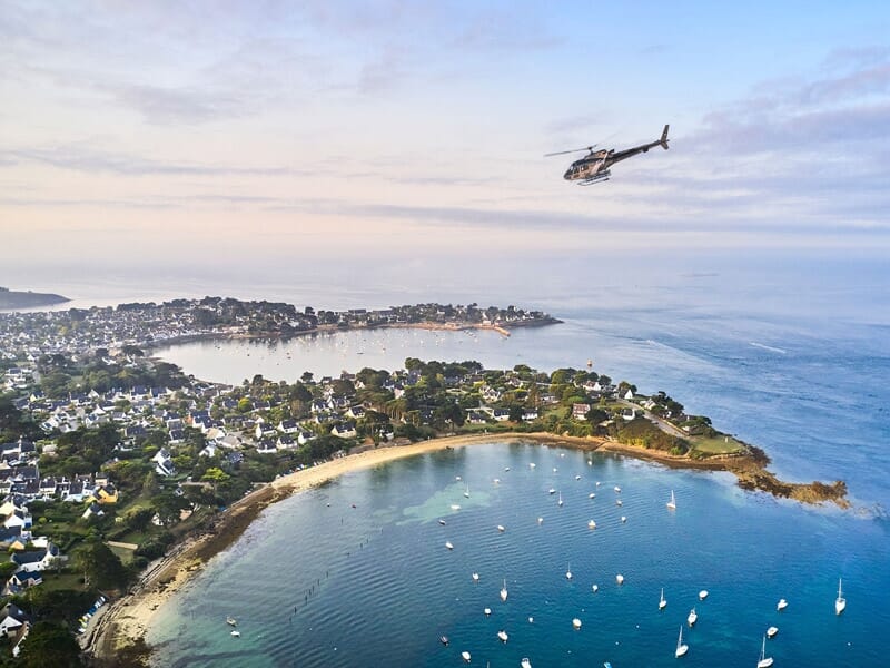 Un hélicoptère volant au-dessus de la côte bretonne du sud au crépuscule, avec un paysage mêlant zones résidentielles et végétation sur fond d'eaux marines calmes.