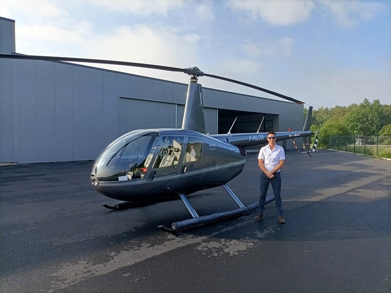 Une personne en chemise blanche et lunettes de soleil debout devant un hélicoptère stationné sur un tarmac, avec de la verdure en arrière-plan et un ciel dégagé au-dessus.