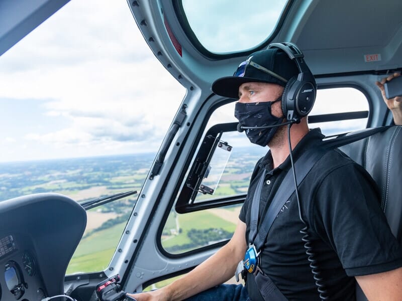 Un pilote habillé en noir, avec un masque facial et un casque, assis dans le cockpit d'un hélicoptère, avec une vue sur la campagne visible à travers la fenêtre.
