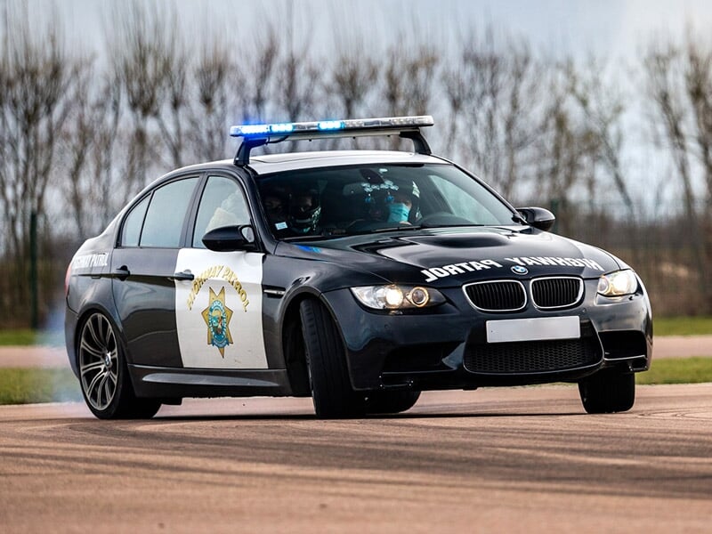 Voiture de police noire avec gyrophares bleus roulant sur une piste en virage, paysage d'arbres en arrière-plan.