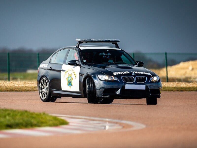 Voiture de police noire en virage sur une piste, avec un conducteur et un passager portant un casque de sécurité.